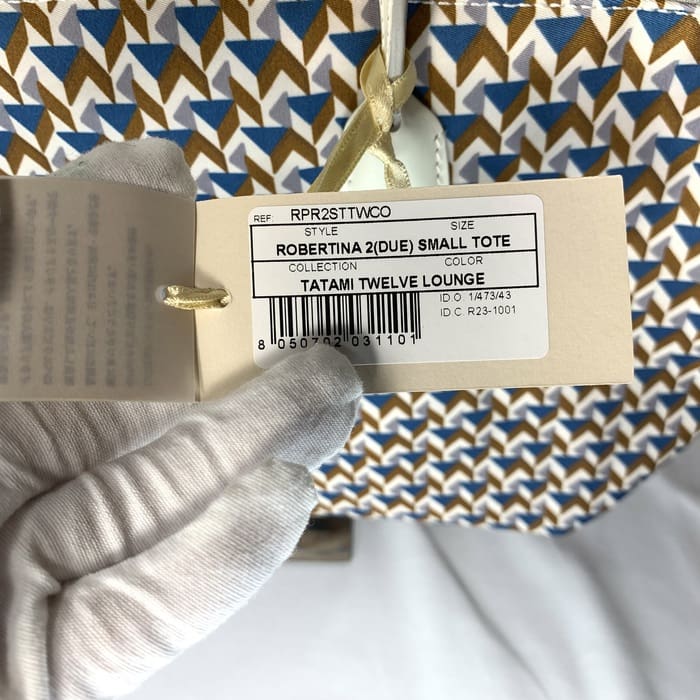 Tangan bersarung tangan memegang label produk "ROBERTINA 2 (DUE) SMALL TOTE" di atas kain bermotif tatami dengan pola geometris berwarna biru, coklat dan putih, yang merupakan gaya khas dari koleksi tas jinjing kecil ROBERTA PIERI.