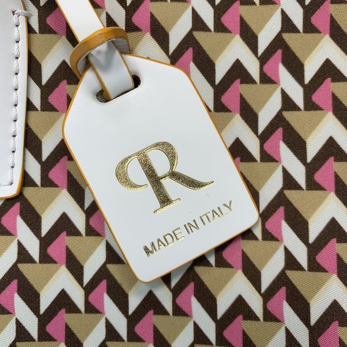 Close-up label bagasi Roberta Pieri putih dengan huruf "R" berwarna emas dan kata-kata "MADE IN ITALY" yang dilekatkan pada tas ransel mini yang menampilkan pola geometris coklat, krem dan merah muda.