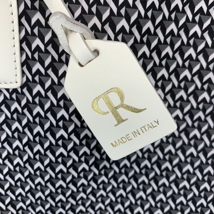 Tag ROBERTA PIERI kulit putih dengan huruf "R" berwarna emas dan kata-kata "MADE IN ITALY" tergantung dari latar belakang kain geometris hitam putih.