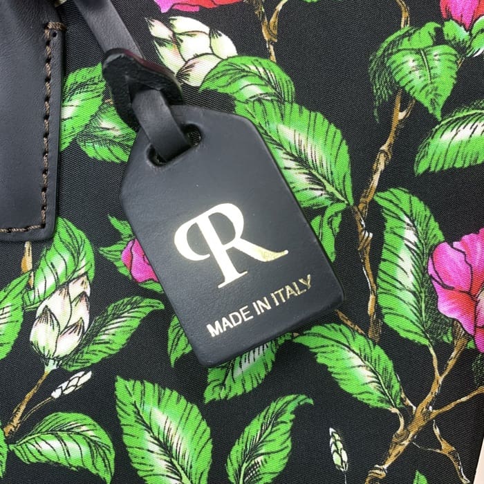 Label bagasi ROBERTA PIERI hitam dengan "R" emas dan teks Italia digantung dari kain bunga dengan daun hijau dan bunga merah muda, melengkapi ransel mini dengan keanggunan Italia yang tak lekang oleh waktu.