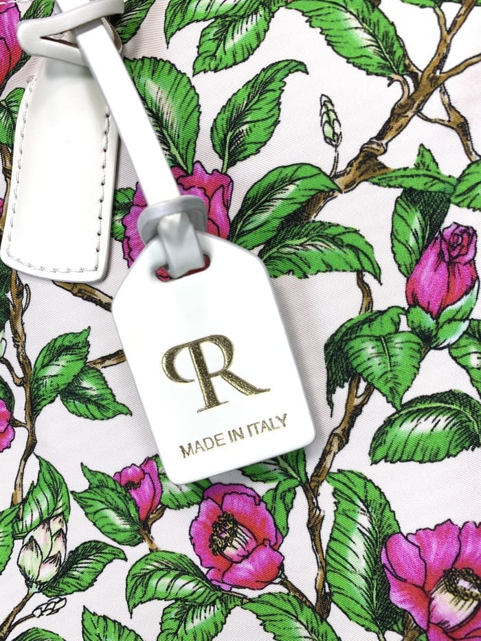 Close-up tas ransel mini ROBERTA PIERI. tangkai dengan daun hijau dan bunga merah muda; Label putih yang melekat pada tas 2 arah menampilkan logo "R" emas dan kata-kata "MADE IN ITALY."