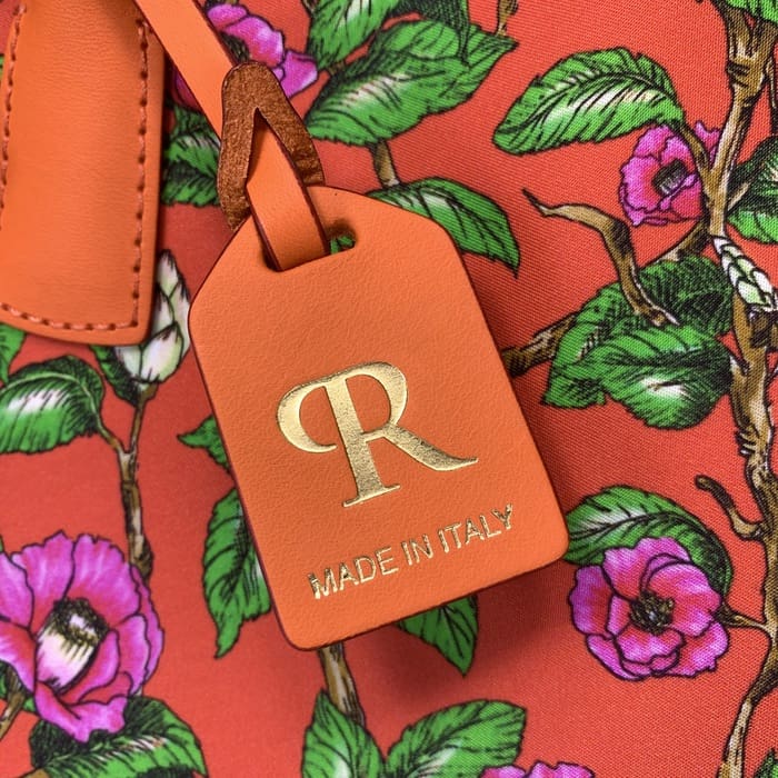 Close-up tag kulit oranye "ROBERTA PIERI" dengan R emas dan teks Italia, dilekatkan pada kain bunga merah dengan bunga merah muda dan daun hijau pada tas ransel mini.